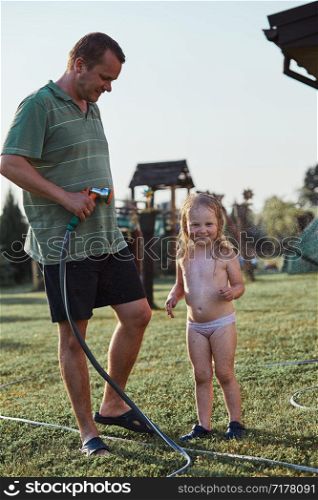 Little cute adorable girl enjoying a cool water sprayed by her father during hot summer day in backyard. Candid people, real moments, authentic situations