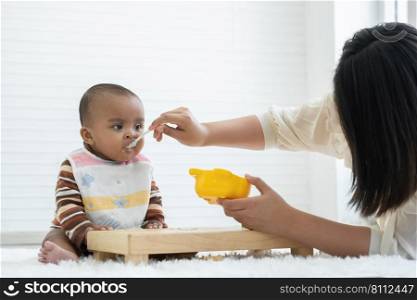 Little cute African newborn baby girl wear apron eating food and sitting on floor at home. Young mother feeding food by spoon to hungry infant daughter. Childcare concept. White background. Copy space