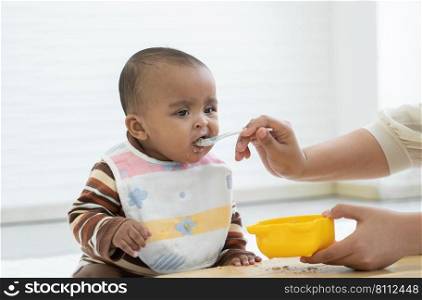 Little cute African newborn baby wear apron eating food and sitting on floor at home. Hand of mother feeding food by spoon to hungry infant daughter. Childcare concept. White background. Copy space