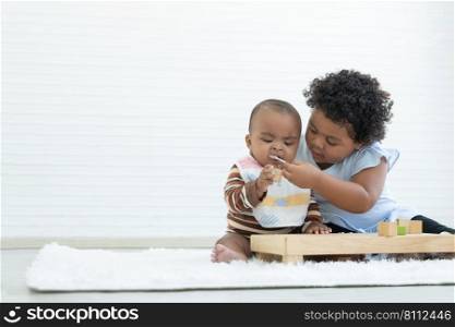 Little cute African older sister try to feeding her newborn sister food while baby playing toys sitting on floor at home. Relationship of Siblings concept. White background. Copy space