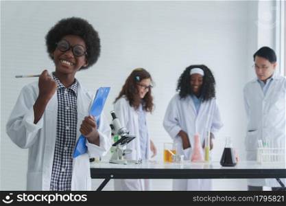 Little cute and smart African boy is taking notes about the science experiment in the laboratory. With his diversity African American Caucasian friends and Asian teacher doing experiment on background
