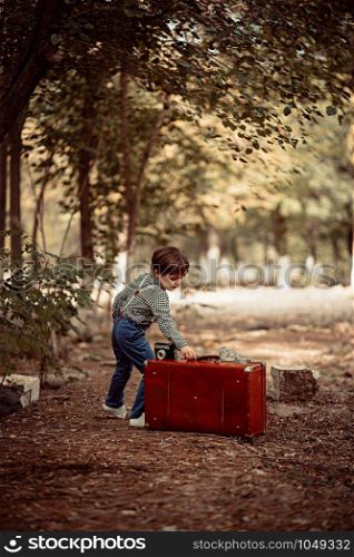 little cute boy in vintage clothes in the Park with vintage suitcase with vintage camera in hand