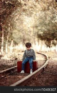 little cute boy in vintage clothes standing on an abandoned railroad with a vintage suitcase in his hands