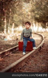 little cute boy in vintage clothes standing on an abandoned railroad with a vintage suitcase in his hands