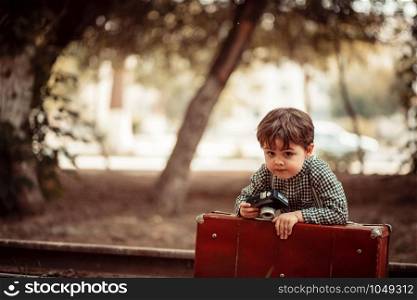 little cute boy in vintage clothes standing on an abandoned railroad with a vintage suitcase with an old camera in his hands