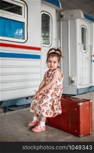 little cute Caucasian girl in vintage clothes with vintage suitcase at small railway station