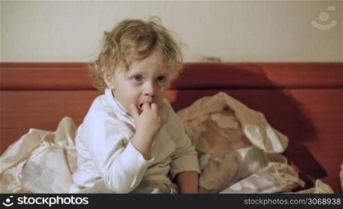 Little cute curly boy watching TV sitting on the bed and putting his fingers into the mouth