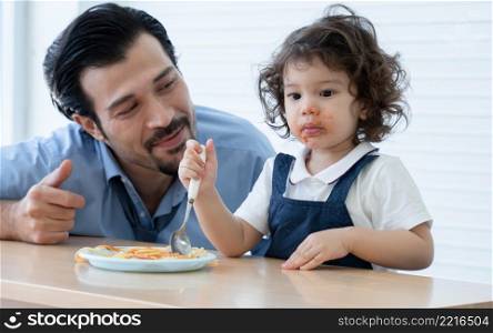 Little cute daughter trying to eat spaghetti with spoon by herself at home and Caucasian young father looking and cheer her up. Adorable kid girl enjoy eating with face is mess up with ketchup