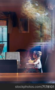 Little cute girl eating a breakfast sitting by the table. Reflections in a glass of window. Candid people, real moments, authentic situations