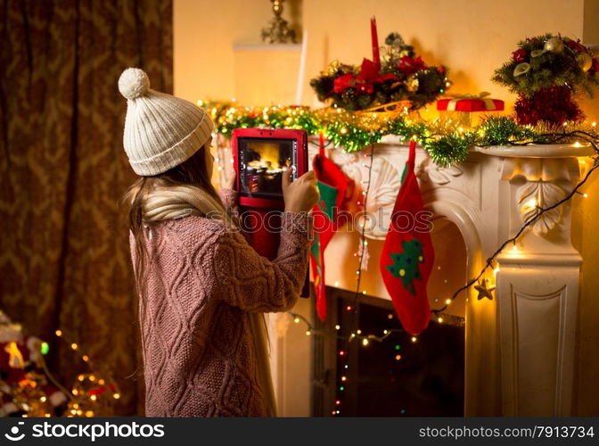 Little cute girl making photo of decorated Christmas fireplace on digital tablet