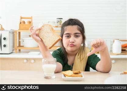 Little cute Indian kid girl dislike breakfast holding sliced bread and thumbs down while having breakfast with a glass of milk, crackers and cookies in kitchen at home