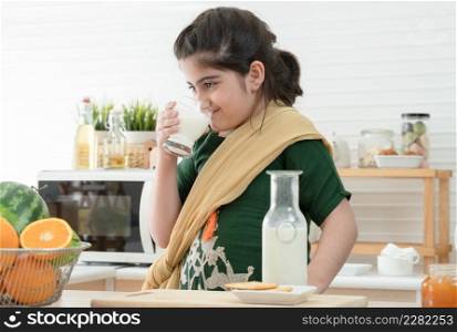 Little cute Indian kid girl holding and drinking a glass of milk for breakfast with bread, jam, crackers and fruits in kitchen at home. A good diet is important to the growth of children concept