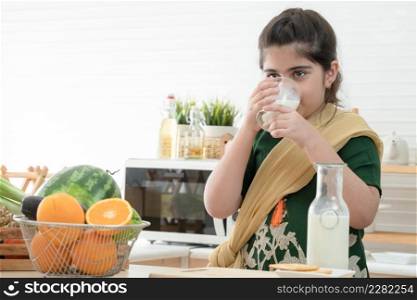 Little cute Indian kid girl holding and drinking a glass of milk for breakfast with crackers and fruits in kitchen at home. A good diet is important to the growth of children concept