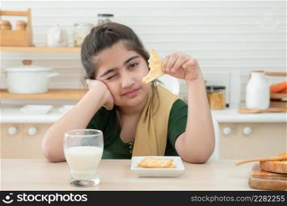 Little cute Indian kid girl holding and eating crackers for breakfast with a glass of milk, jam and bread in kitchen at home. A good diet is important to the growth of children concept