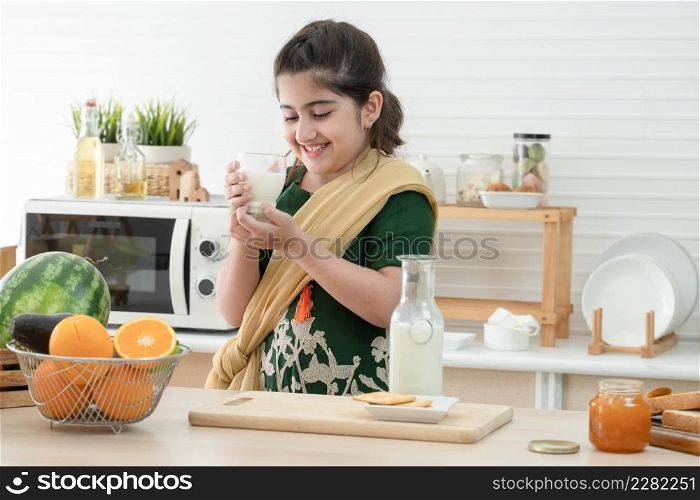 Little cute Indian kid girl smiling and holding a glass of milk for breakfast with bread, jam, crackers and fruits in kitchen at home. A good diet is important to the growth of children concept