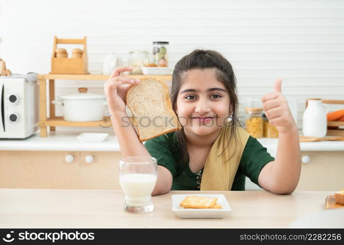 Little cute Indian kid girl smiling holding sliced bread and thumbs up while having breakfast with a glass of milk and crackers in kitchen at home. A good diet is important to the growth of children