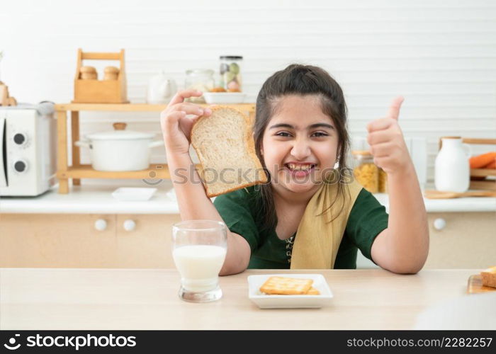Little cute Indian kid girl smiling holding sliced bread and thumbs up while having breakfast with a glass of milk and crackers in kitchen at home. A good diet is important to the growth of children