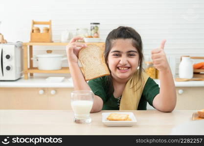 Little cute Indian kid girl smiling holding sliced bread and thumbs up while having breakfast with a glass of milk and crackers in kitchen at home. A good diet is important to the growth of children