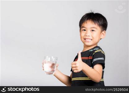 Little cute kid boy 5-6 years old smile drinking fresh water from glass and show thumb up finger for good sign in studio shot isolated on white background, Asian children preschool, Daily life health
