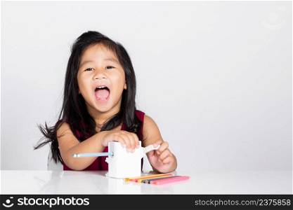 Little cute kid girl 3-4 years old smile using pencil sharpener while doing homework in studio shot isolated on white background, Asian children preschool sharpening color pencils, education concept