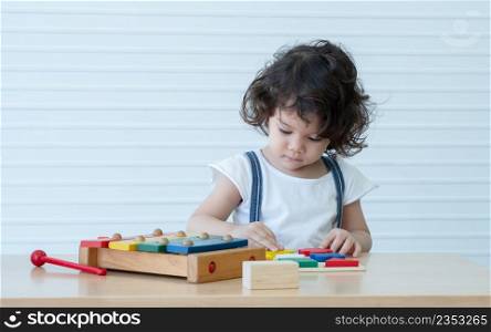 Little cute mixed race kid girl, Caucasian Asian, is concentrating on playing with colorful wooden puzzle pieces blocks and xylophone toy on table. White background. Copy space
