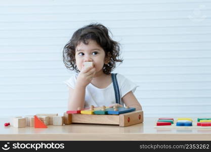 Little cute mixed race kid girl, Caucasian Asian, playing toys build a wooden block, xylophone and colorful puzzle piece on table. White background. Copy space