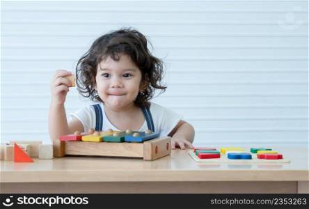 Little cute mixed race kid girl, Caucasian Asian, smiling and playing toys build a wooden block, xylophone and colorful puzzle piece on table. White background. Copy space