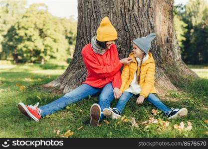 Little daughter and her mother have fun together, dressed warm, sit near big tree on green grass, look at each other with love. Affectionate mom and small female kid have good mood, spend weekends