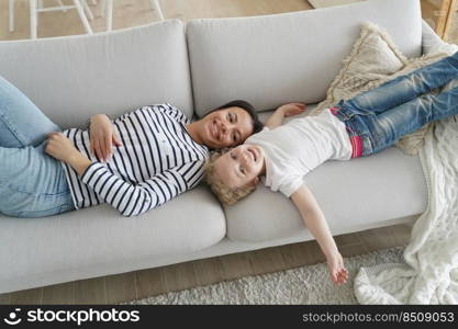 Little daughter lying on comfortable sofa with mother, smiling looking at camera. Mom and kid girl relaxing have lazy weekend together, enjoy comfort rest. Happy motherhood concept.. Little daughter and mother lying on sofa relax, have lazy weekend together. Happy motherhood