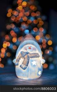 Little decorative snowman toy on the table over glowing Christmas tree background, happy winter holidays, traditional New Year and winter character