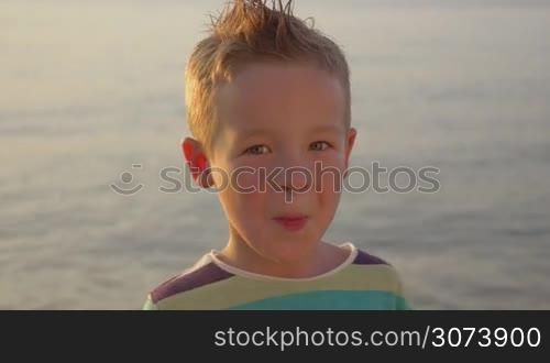 Little funny boy is making faces in front of the camera, sea is on the background.