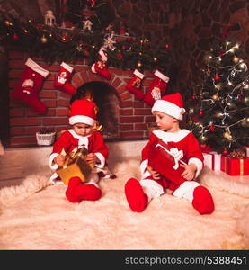 Little girl and boy are sitting near fireplace and christmas tree with gift boxes. Brother and sister in santa suits