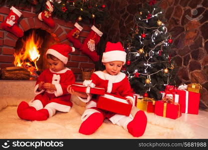 Little girl and boy are sitting near fireplace and christmas tree with gift boxes. Brother and sister in santa suits