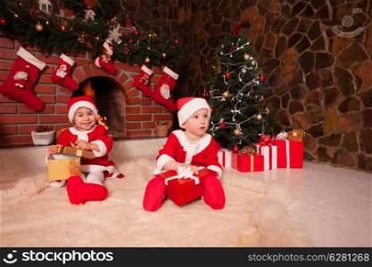 Little girl and boy are sitting near fireplace and christmas tree with gift boxes. Brother and sister in santa suits
