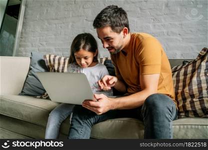 Little girl and her father having fun and using a laptop together while sitting on a couch at home. Monoparental family concept.