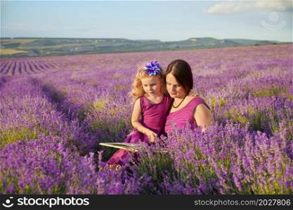 Little girl and mother at meadow of lavender reading a book. Family care and nature composition.