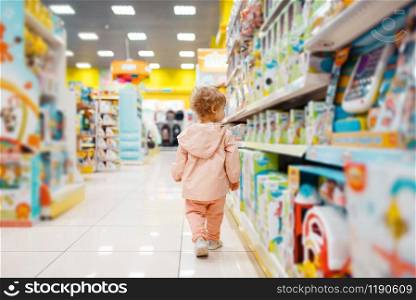 Little girl at the shelf choosing toys in kids store, side view. Daughter in supermarket, family shopping, young customer. Little girl choosing toys in kids store