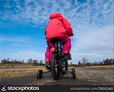 Little girl biker on gravle road with supporting wheels on gravel road at spring with blue sky