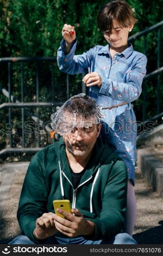 Little girl blowing up a water-filled balloon over her father&rsquo;s head to get him to stop staring at his smartphone.. Girl blowing up a water-filled balloon over her father&rsquo;s head