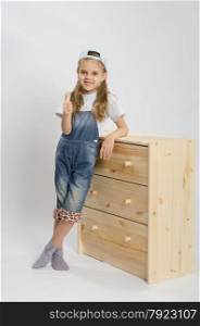 Little girl - collector of furniture in overalls relies on a chest of drawers and shows class. Girl in overalls collector of furniture based on the chest shows class