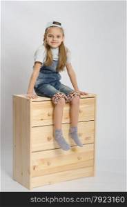 Little girl - collector of furniture in overalls relies on a chest of drawers and shows class. Girl in overalls collector relies on the chest shows class