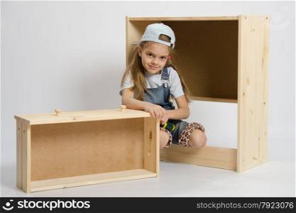 Little girl - collector of furniture sitting around the wooden parts of the chest. Little girl in overalls sits beside the garbage furniture elements Chest