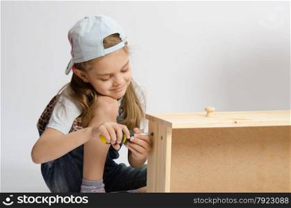 Little girl - collector of furniture twists the screw with a screwdriver in the wooden frame of the chest. Little girl in overalls collector of furniture screws screwdriver screw
