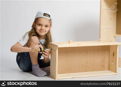 Little girl - collector of furniture twists the screw with a screwdriver in the wooden frame of the chest. Little girl in overalls collector collects furniture drawer of the dresser