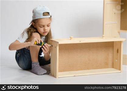 Little girl - collector of furniture twists the screw with a screwdriver in the wooden frame of the chest. Little girl in overalls collector of furniture making box on the dresser