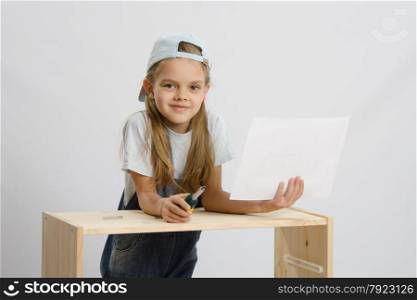 Little girl - collector of furniture with a screwdriver and instructions for assembly in the hands collects wooden cabinet. Girl-collector of furniture with a screwdriver and drawing collects furniture