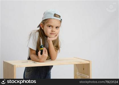 Little girl - collector of furniture with a screwdriver in his hand thoughtfully leaned on unfinished furniture and laid her head on her hand. Girl-collector of furniture with a screwdriver wearily leaned on the cabinet