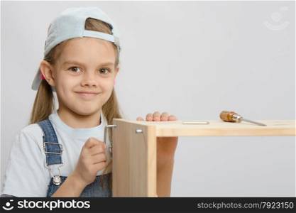Little girl - collector of furniture with an Allen key is trying to screw a screw into the wooden frame of the chest. Girl in overalls furniture collector tries to tighten the screw