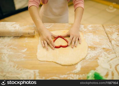 Little girl cook in apron makes cookies in the shape of a heart on the kitchen. Kid cooking pastry, child chef preparing cake