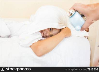 Little girl covering head with pillow and being awakened by alarm clock